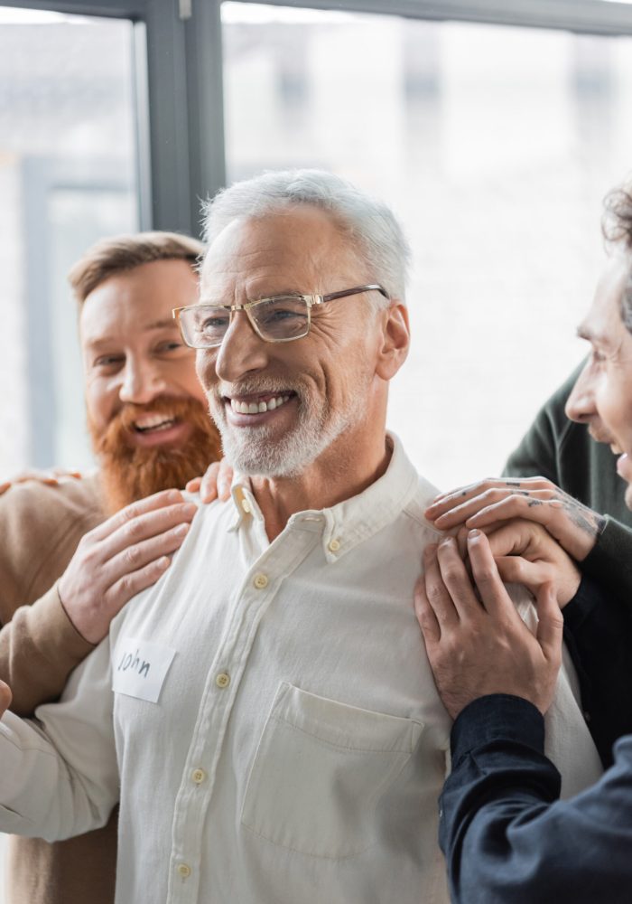 Positive people hugging middle aged man with alcohol addiction during group therapy in rehab center,stock image
