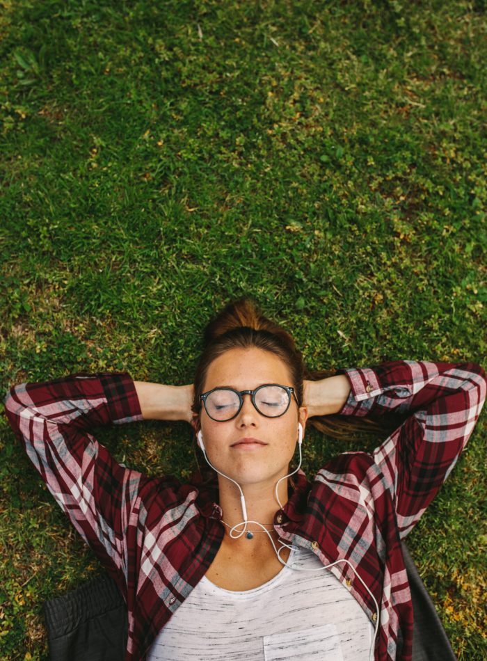 Top view of a woman lying on grass with hands behind head. Female with closed eyes relaxing on grass and listening to music using earphones.